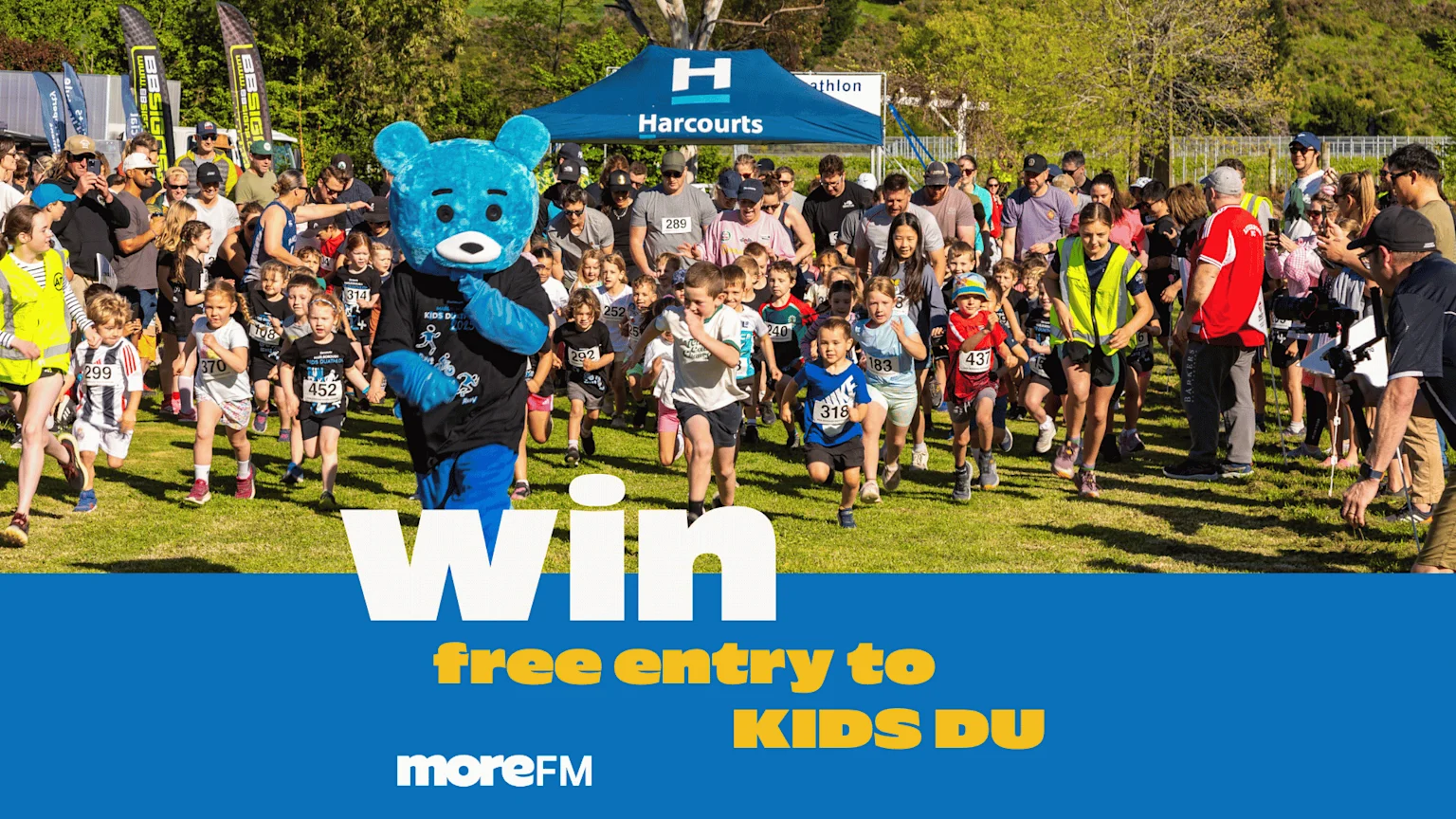 A blue mascot bear and many children run a race on a grassy field, with adults watching, a "Harcourts" tent, and "Win free entry to KIDS DU" text.