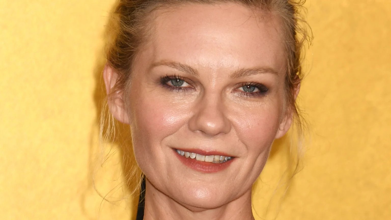 Close-up of a fair-skinned woman with blonde hair pulled back, light blue eyes, and reddish-brown lips, smiling slightly to reveal a small gap between her upper front teeth, against a yellow background.