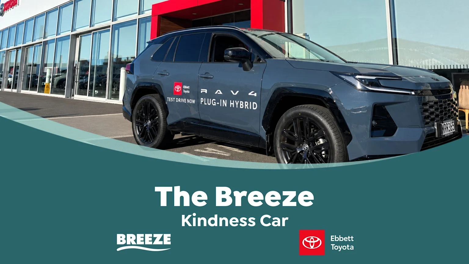 A grey Toyota RAV4 Plug-in Hybrid "The Breeze Kindness Car" with "Ebbett Toyota" branding is parked in front of a dealership, with "TEST DRIVE NOW" text visible on its side.