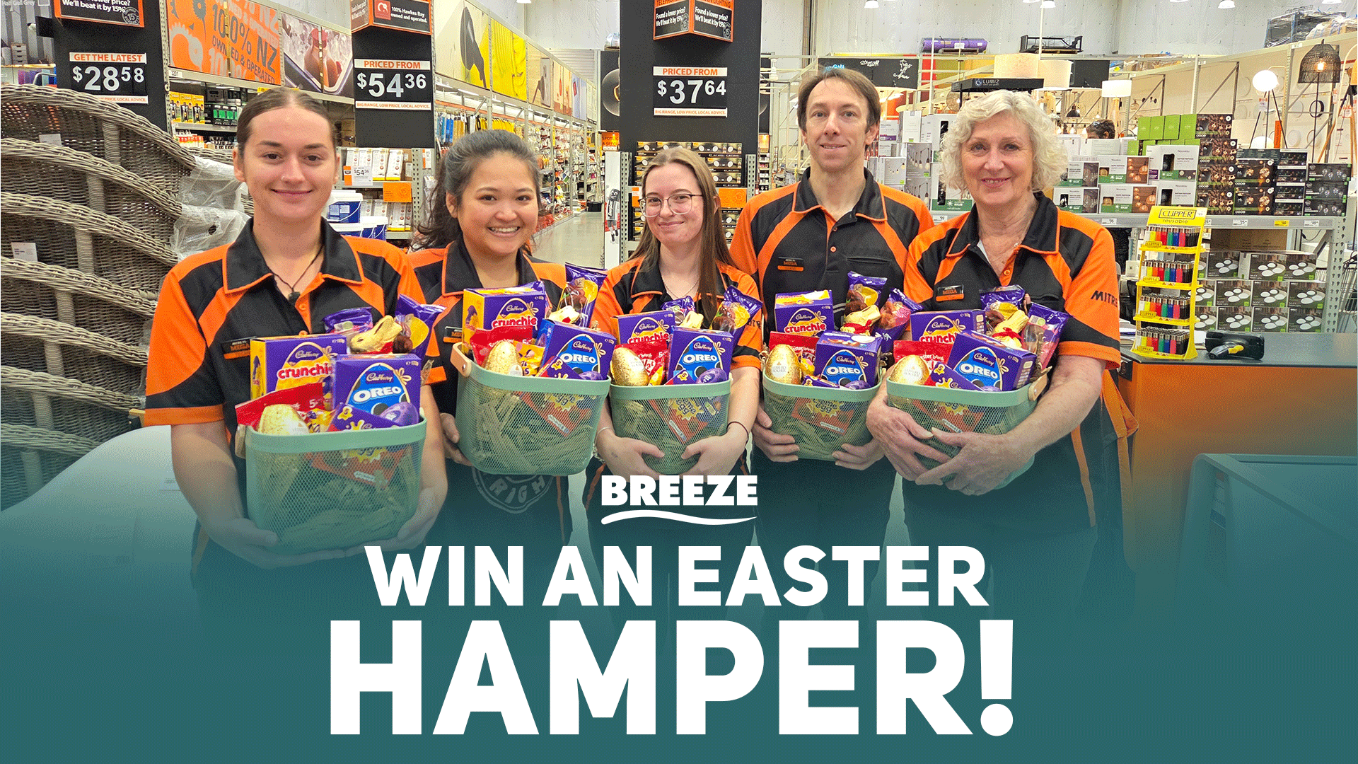 Five smiling Mitre 10 employees in orange and black uniforms hold green baskets filled with Cadbury Easter chocolates, promoting an Easter hamper win in a retail store.
