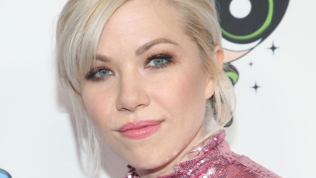 Close-up of Carly Rae Jepsen with blonde hair and blue eyes, wearing a pink sequined top, smiling slightly against a white background with a subtle cartoon graphic.