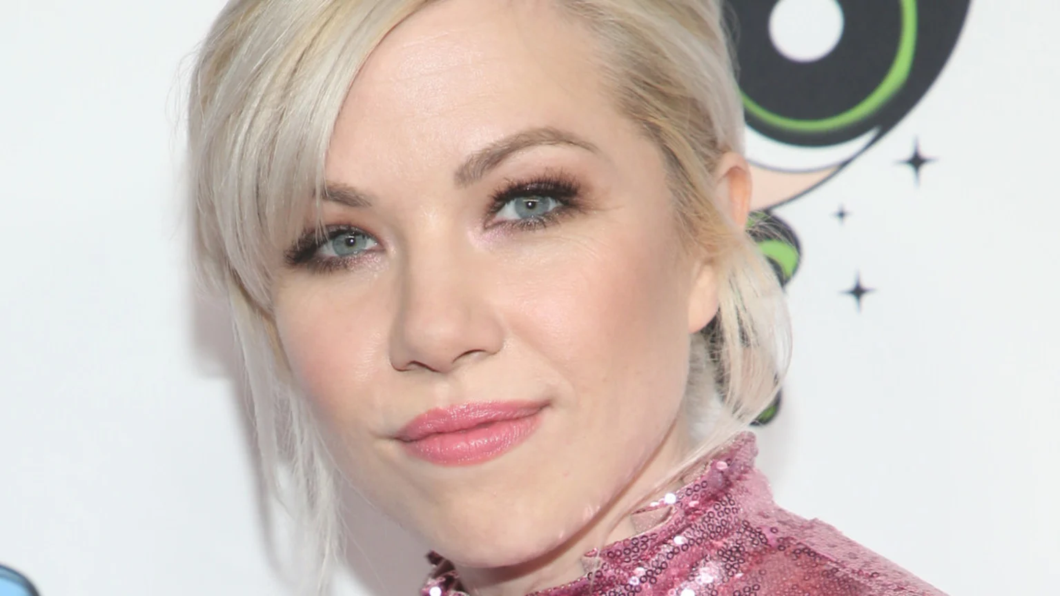 Close-up of Carly Rae Jepsen with blonde hair and blue eyes, wearing a pink sequined top, smiling slightly against a white background with a subtle cartoon graphic.