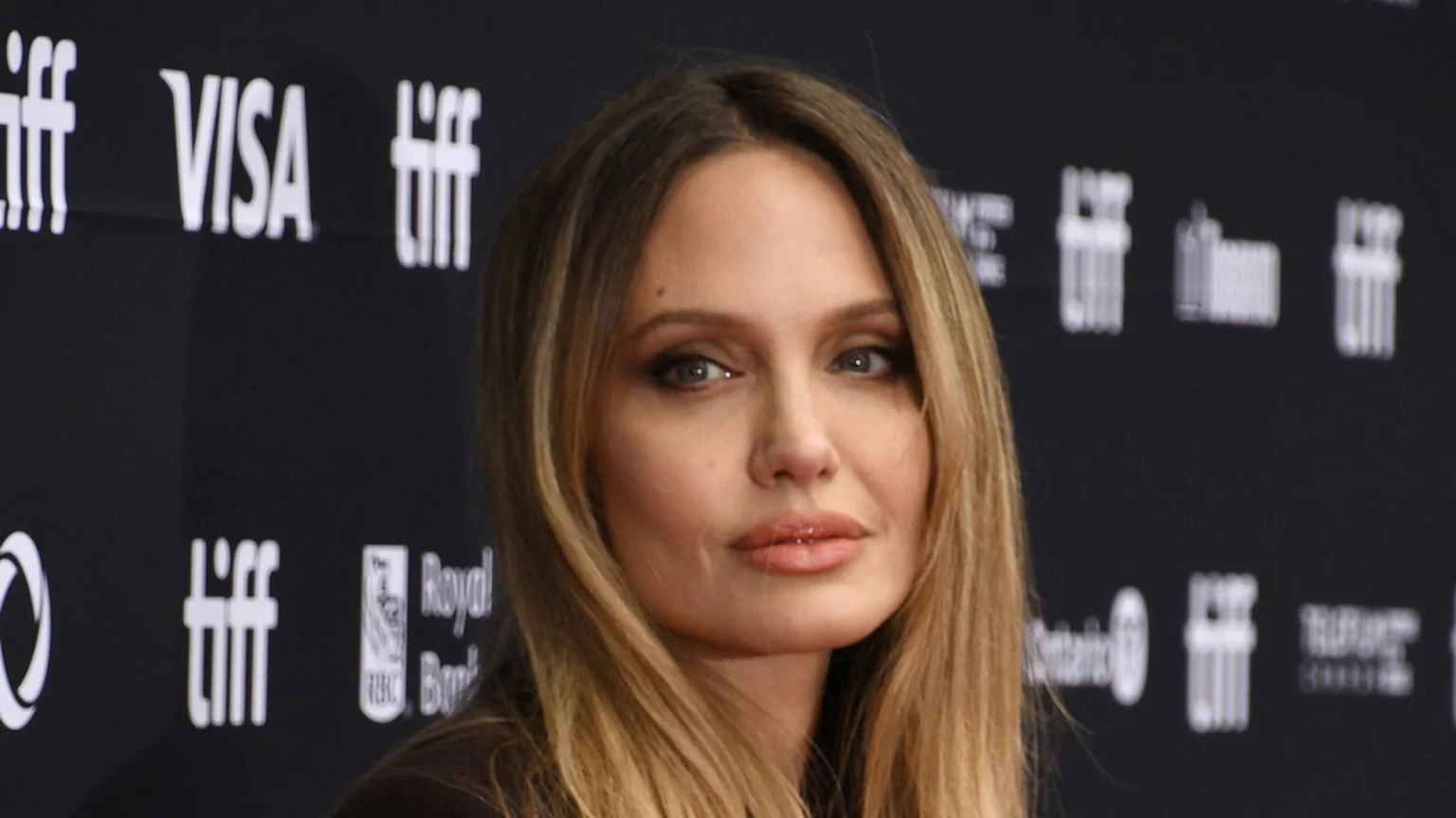 A close-up of Angelina Jolie looking directly at the viewer, with long, light brown hair and soft makeup, against a dark background with white event logos.