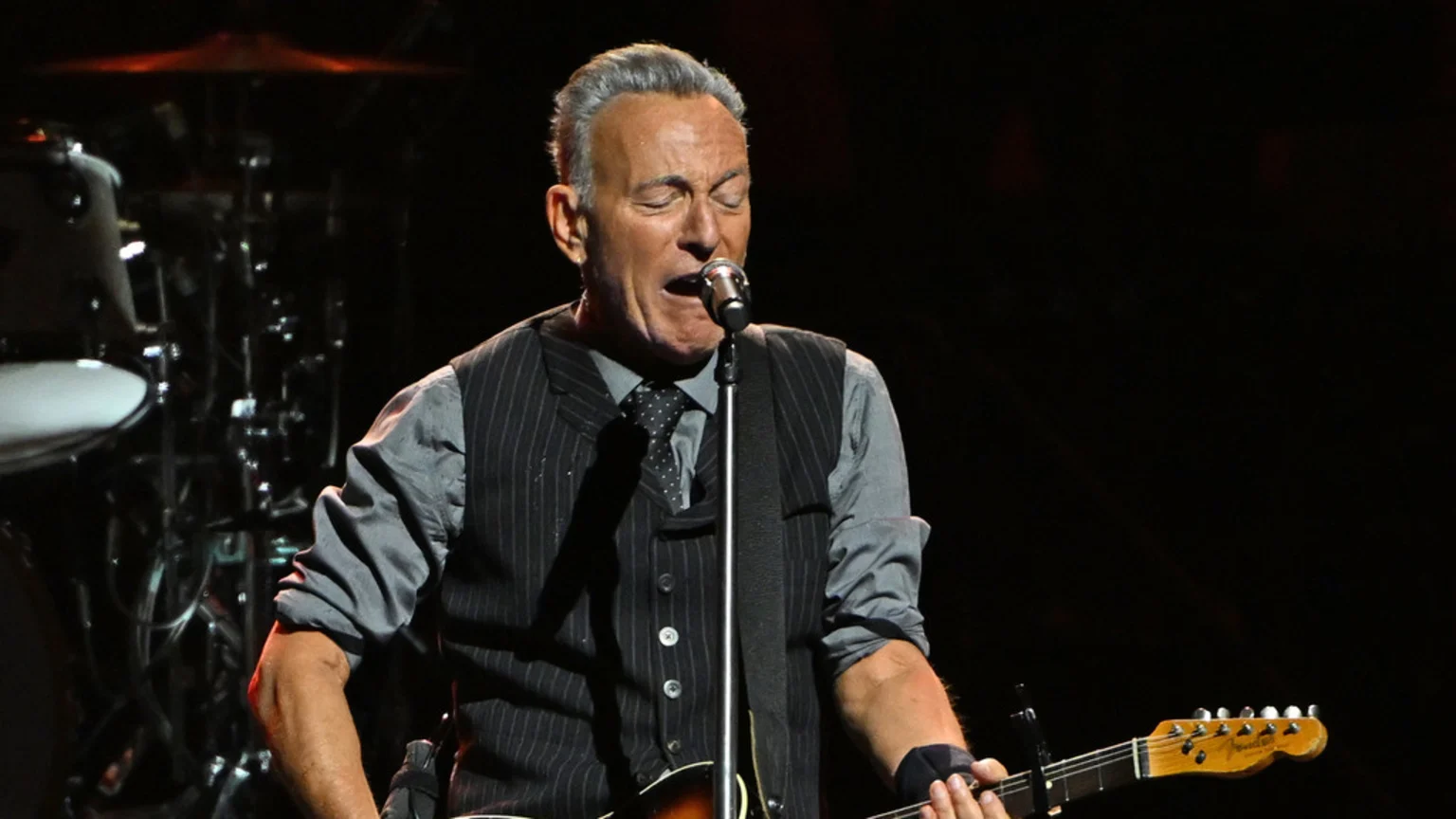 Bruce Springsteen sings into a microphone while playing an electric guitar on a dark stage, wearing a pinstripe vest and grey shirt.