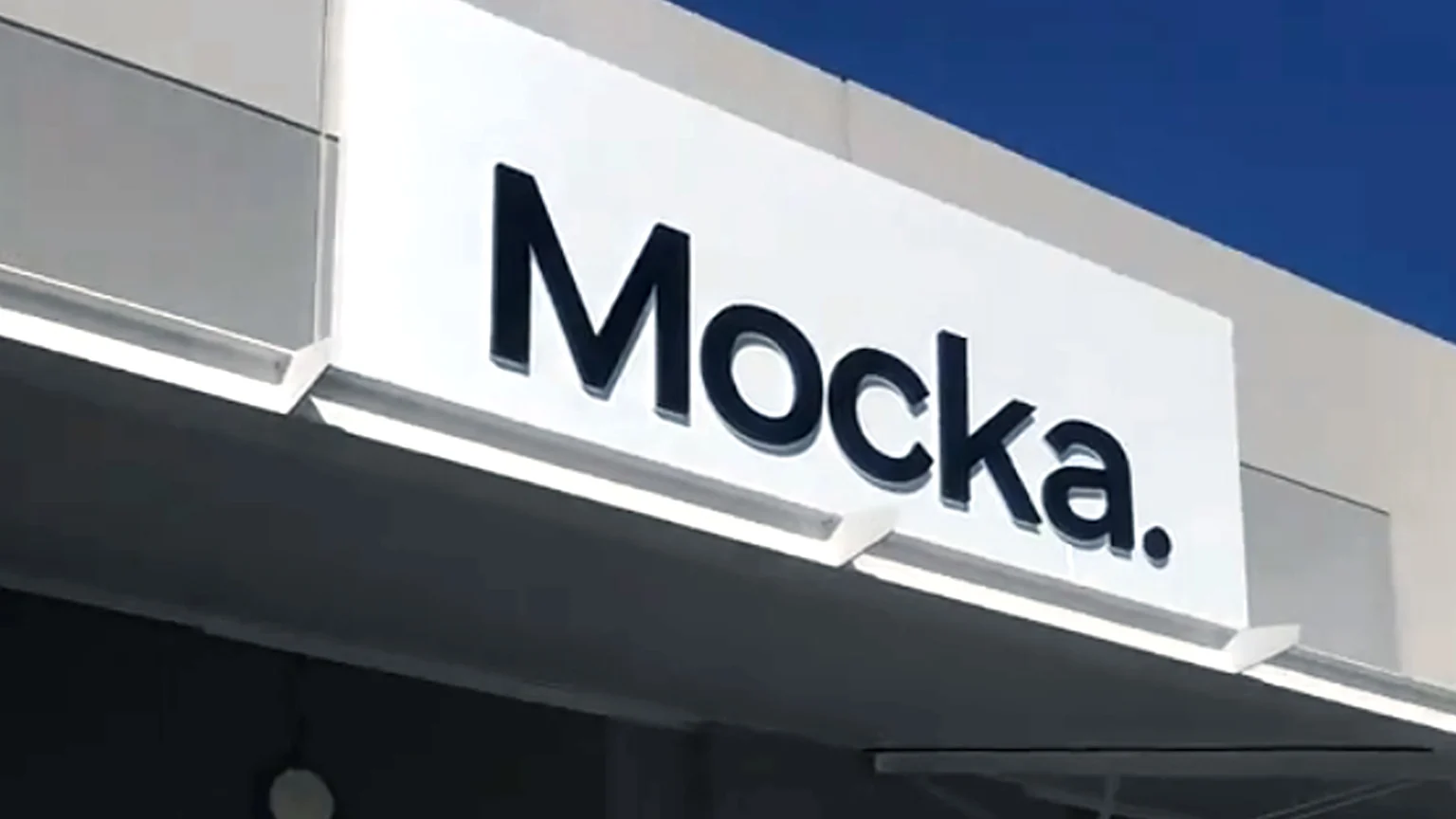 A white store sign with the black, raised letters "Mocka." illuminated from below, attached to a grey building facade against a blue sky.