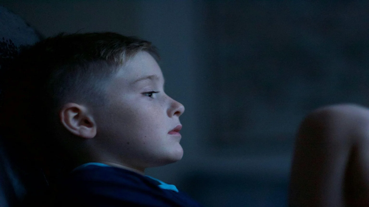 A young boy with freckles and a short haircut is shown in profile, looking intently to the right with a serious expression in a dimly lit setting.