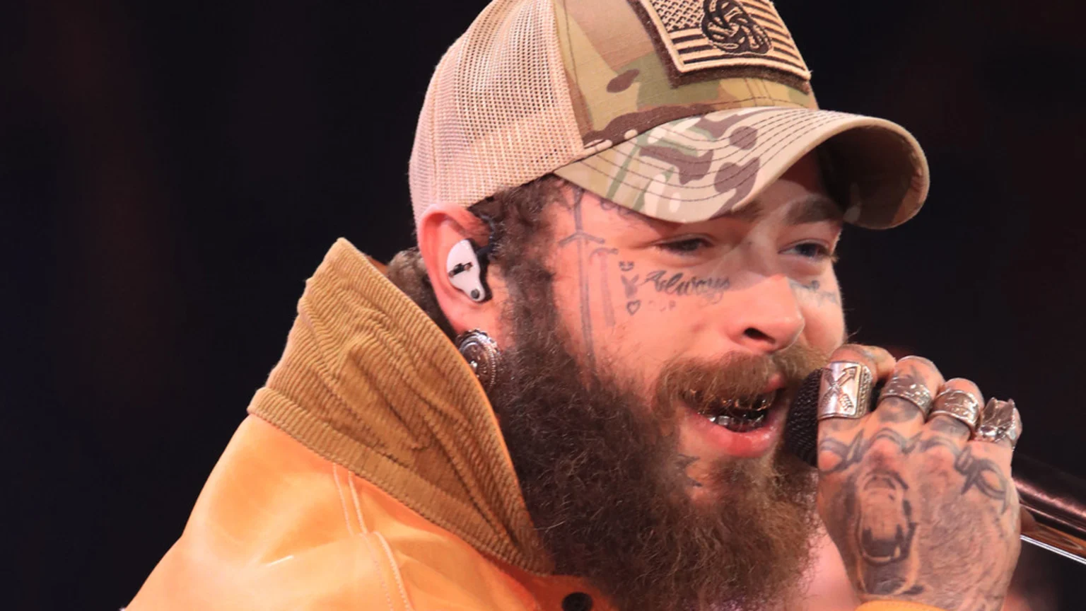 Close-up of Post Malone, a man with a beard, face tattoos, and grills, wearing a camouflage hat and orange jacket, holding a microphone.