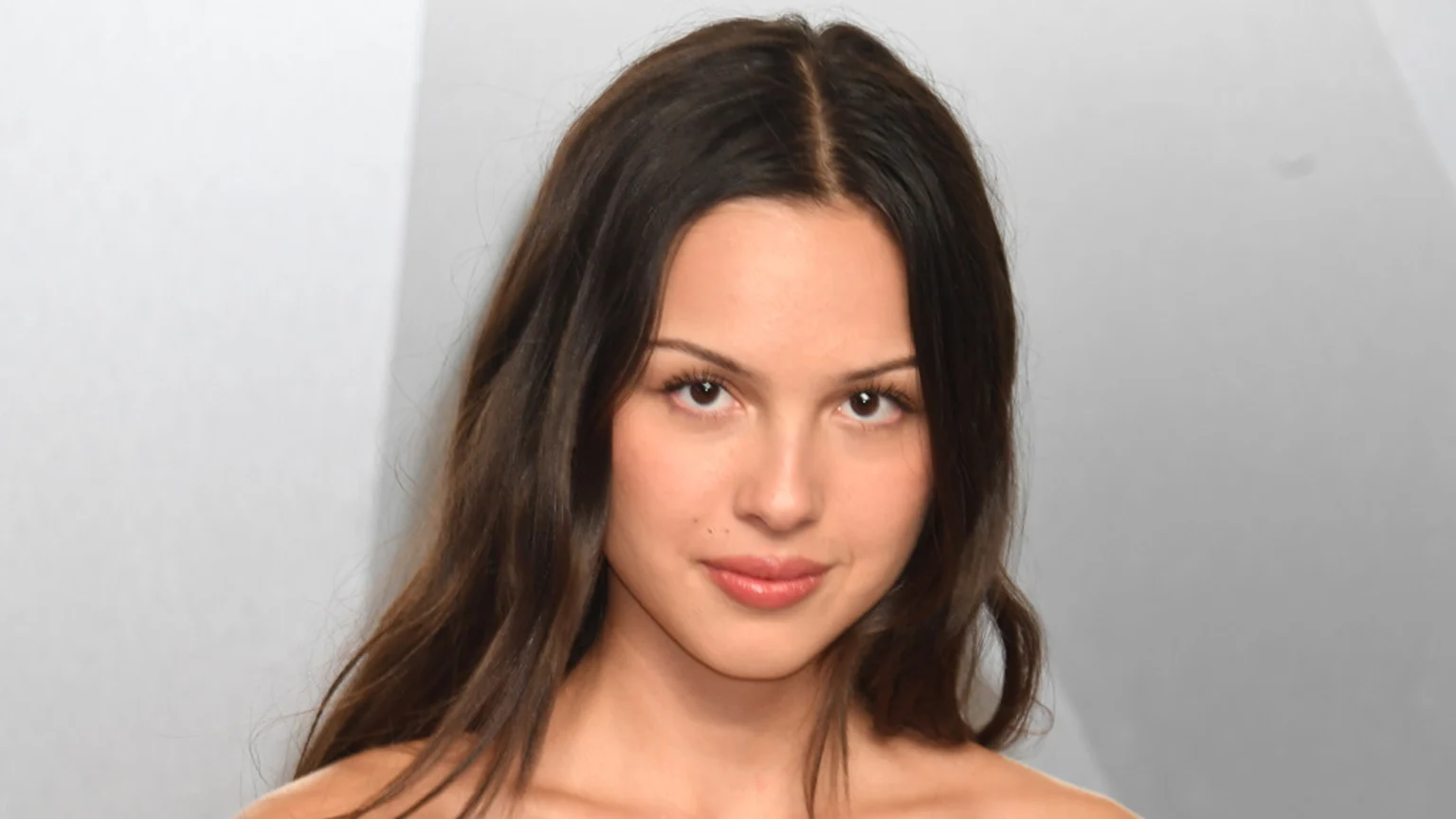 A young woman with long dark hair parted in the middle, light makeup, and a gentle smile looks directly ahead against a plain light gray background.