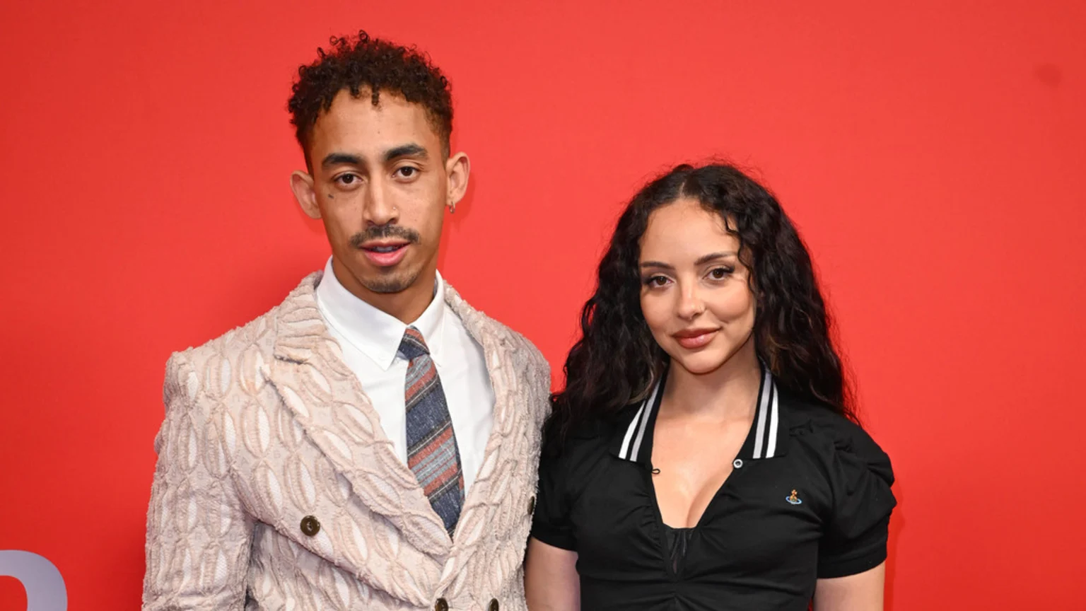 A man with curly hair, a nose ring, and an ear piercing, wearing a textured suit, stands with a woman with long, dark curly hair, wearing a black top with a small orb emblem, against a red background.