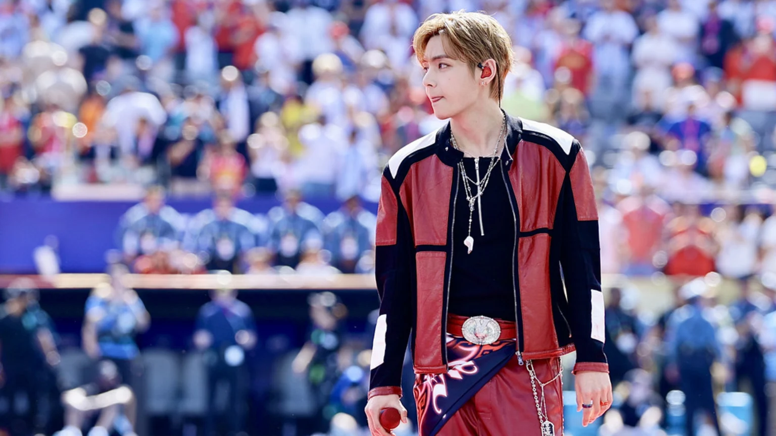 A person with light brown hair, wearing a red and black jacket and red pants, looks right with their tongue out, holding a red microphone in a blurred stadium.