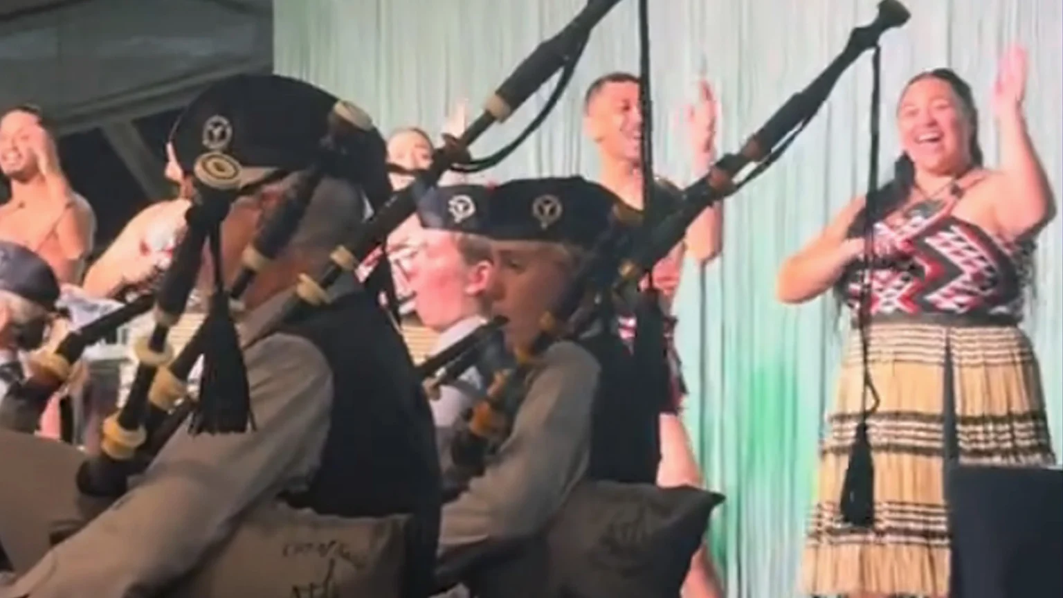 Two bagpipers in uniform play in the foreground while smiling people, including a woman in a patterned top and woven skirt, clap in the background against a green curtain.