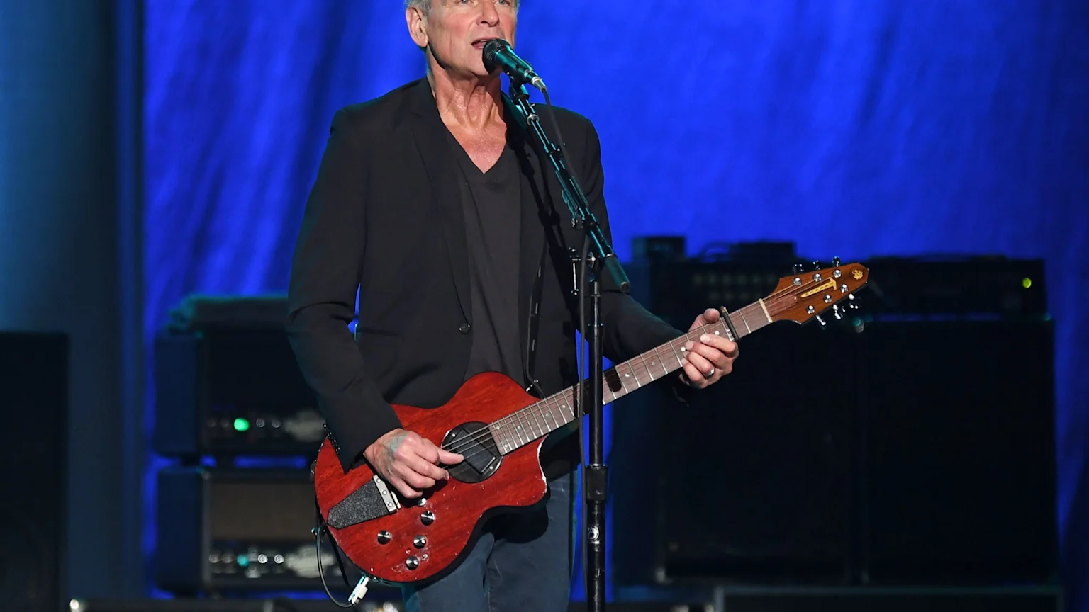 Lindsey Buckingham, a man with gray hair, sings into a microphone while playing a red-brown guitar on stage, with a blue curtain background.