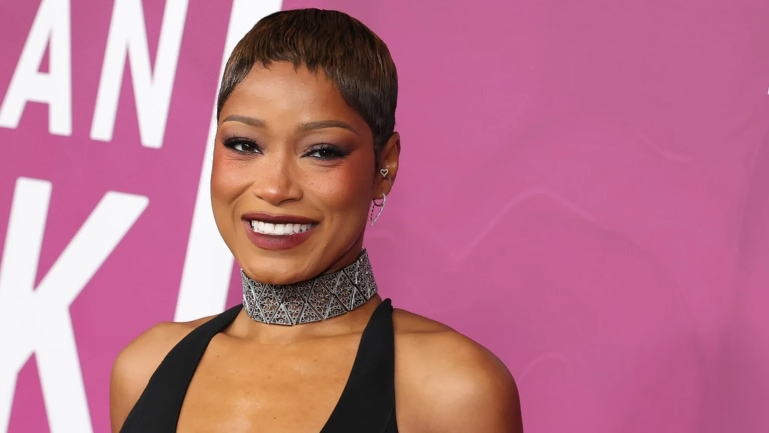 A smiling Keke Palmer with short brown hair, freckles, and dark eye makeup wears a black top, a silver patterned choker necklace, and heart-shaped earrings against a pink background.