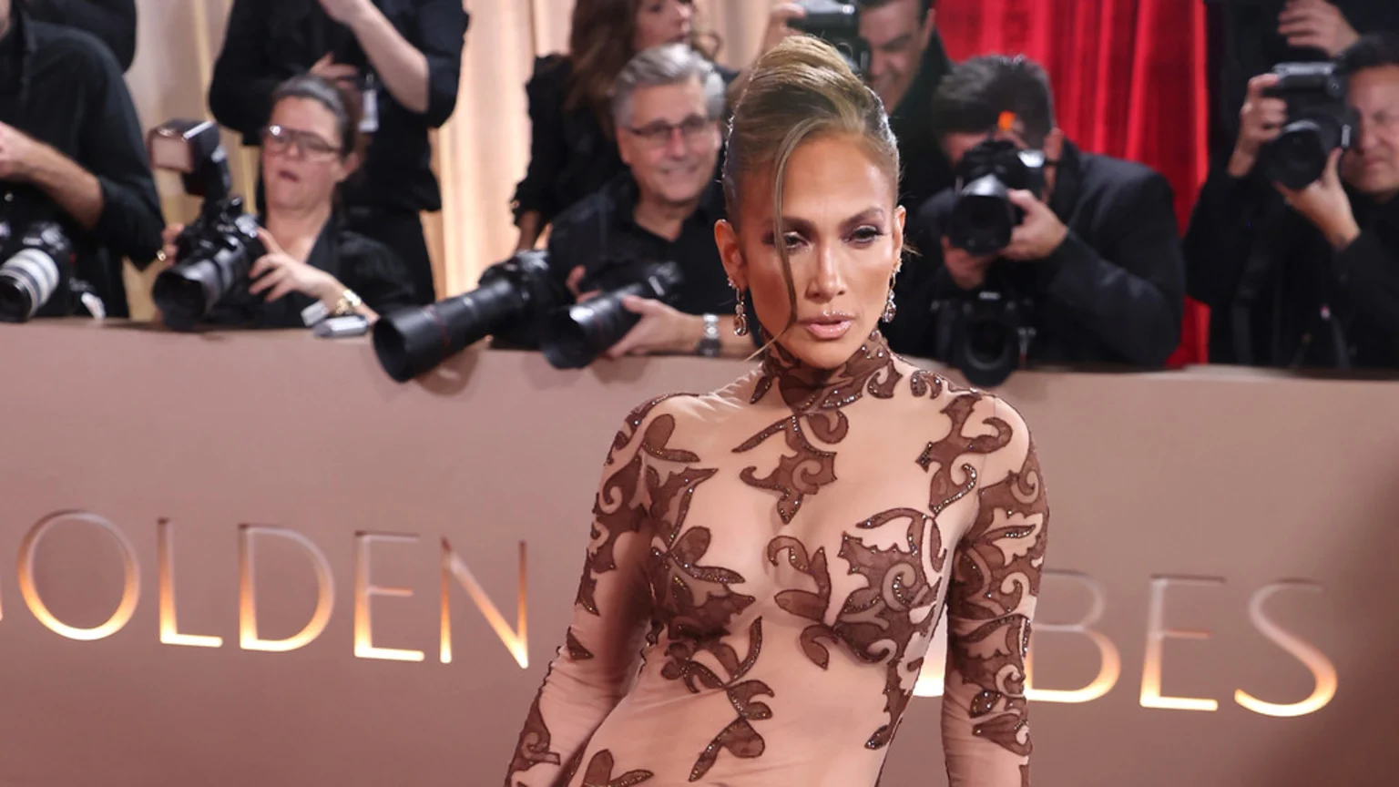 Jennifer Lopez poses in a sheer brown patterned dress with intricate designs, with photographers behind her and a "Golden Globes" sign visible.