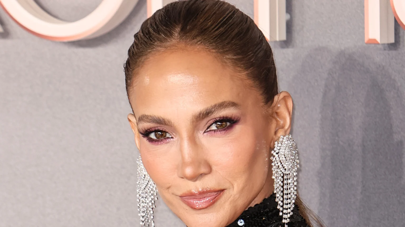 Close-up of Jennifer Lopez with her hair pulled back, wearing purple eyeshadow, long lashes, glossy lips, and large, sparkly chandelier earrings, smiling slightly.