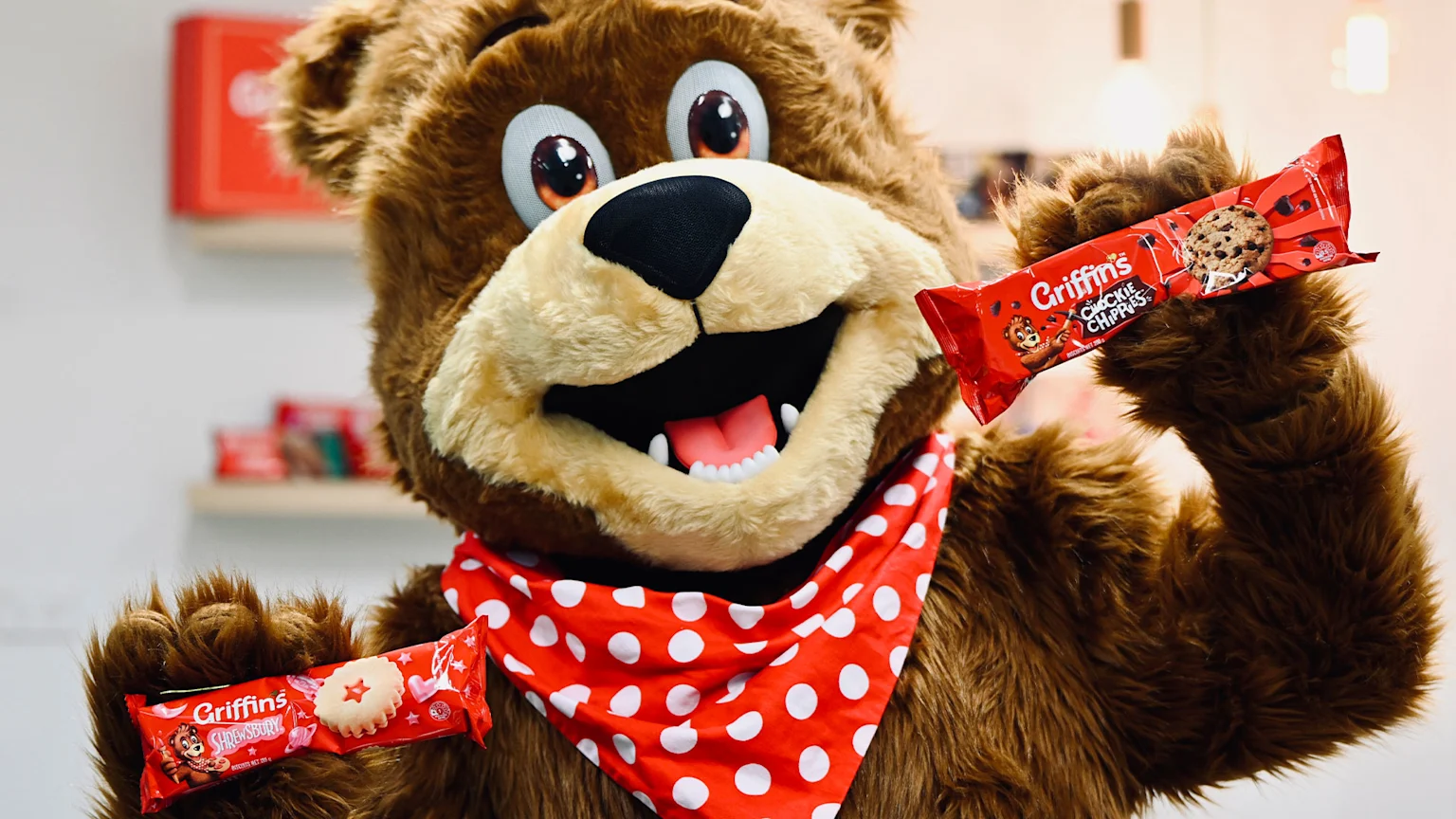A cheerful brown bear mascot wearing a red polka dot bandana holds two red Griffin's biscuit packages labeled 'Chockie Chippies' and 'Shrewsbury' against a blurred indoor backdrop with shelves and warm lighting.