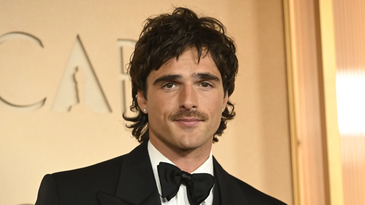 Jacob Elordi, with wavy dark hair and a mustache, wears a tuxedo with a bow tie, looking directly at the camera against a blurred tan background with part of an 'OSCAR' logo.