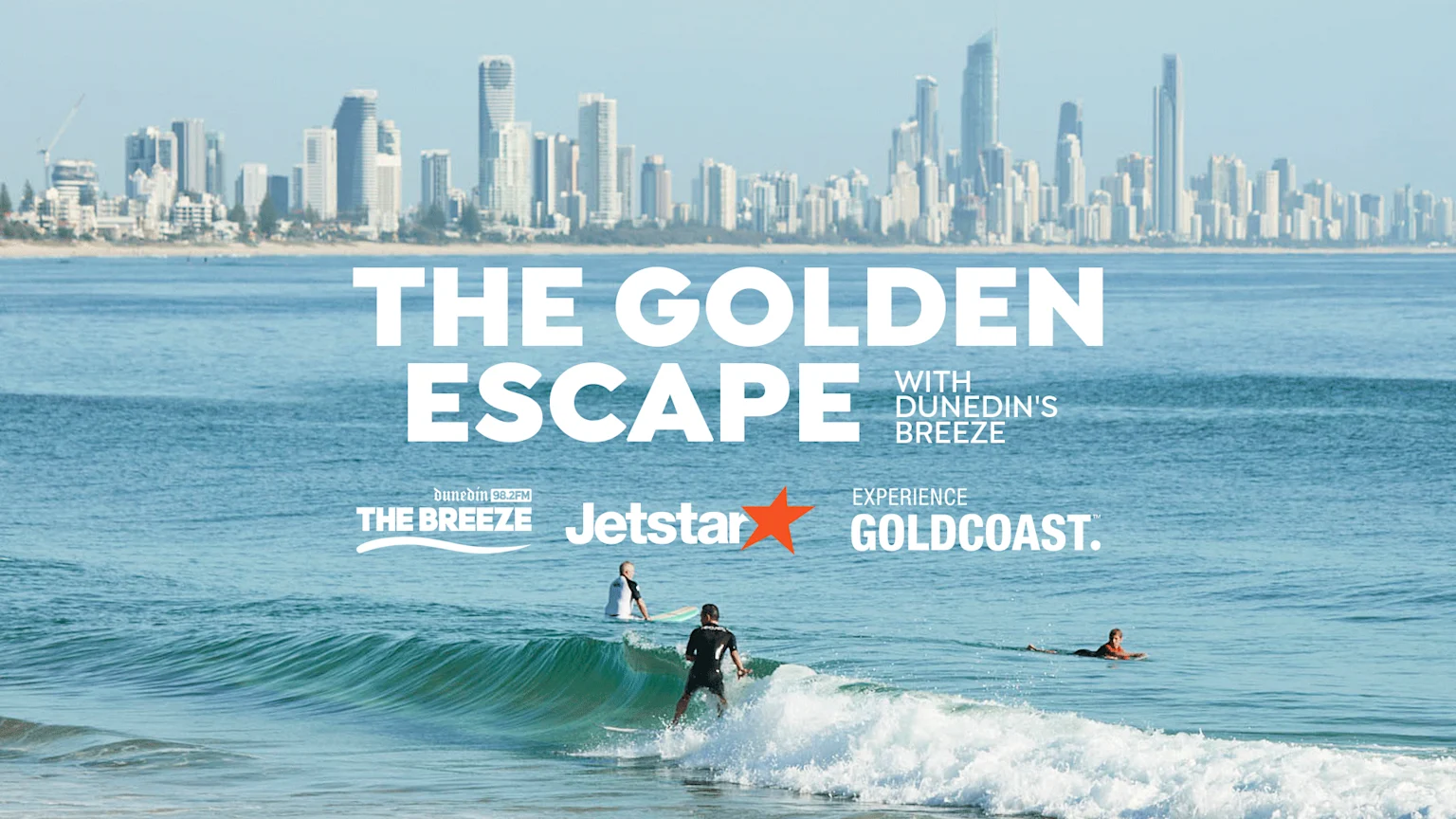 A sunny beach scene with surfers and a city skyline in the background, overlaid with white text "THE GOLDEN ESCAPE WITH DUNEDIN'S BREEZE" and sponsor logos at the bottom.
