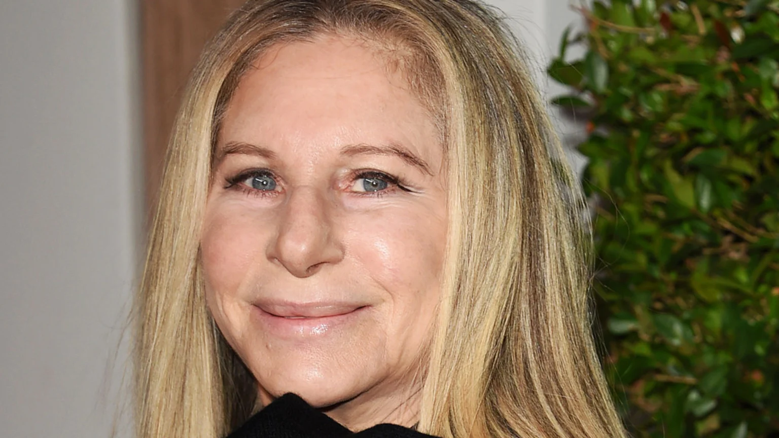 Close-up portrait of Barbra Streisand smiling, with blonde hair, blue eyes, and a blurred background featuring a light wall and green foliage.