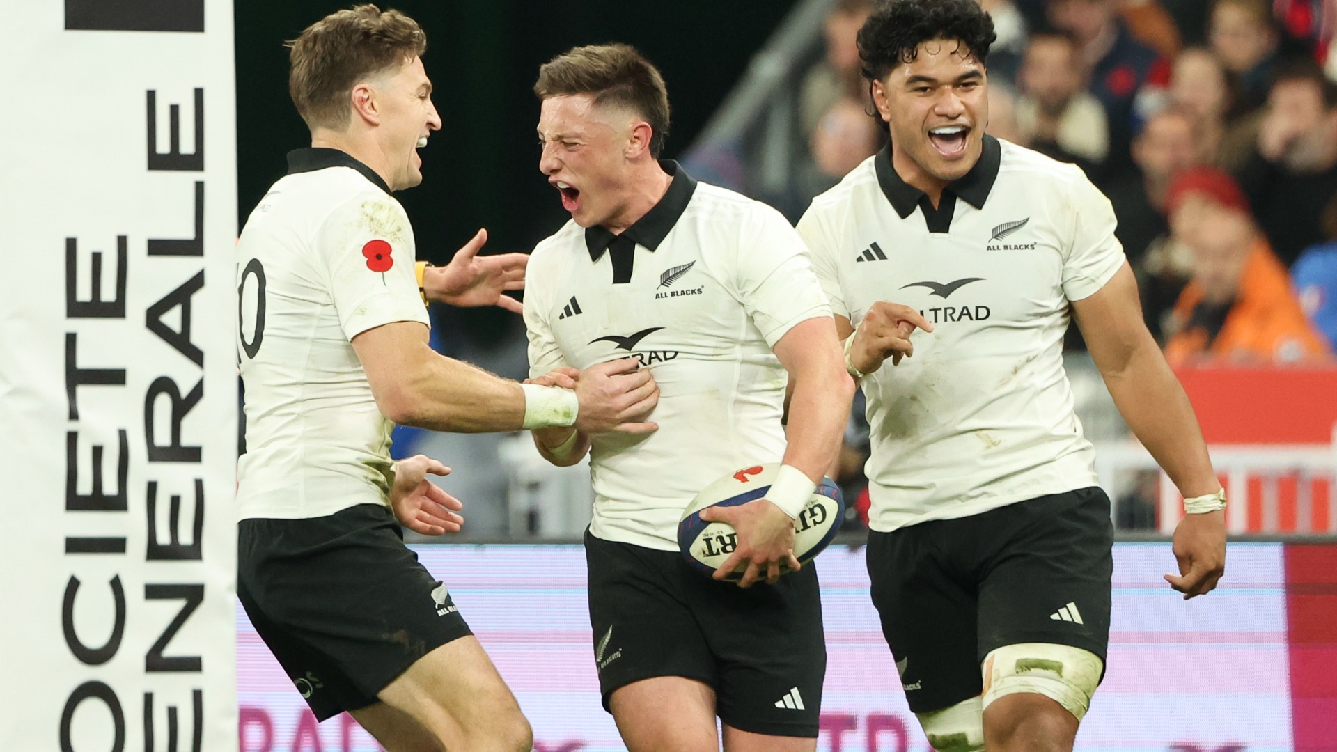 Three All Blacks rugby players celebrate excitedly on the field; the central player shouts while holding a rugby ball, flanked by two smiling teammates.