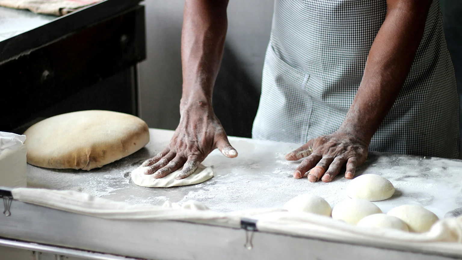 A person with floured hands, wearing a checkered apron, is pressing a small piece of dough on a flour-dusted surface, with a large dough mass and several smaller dough balls nearby.