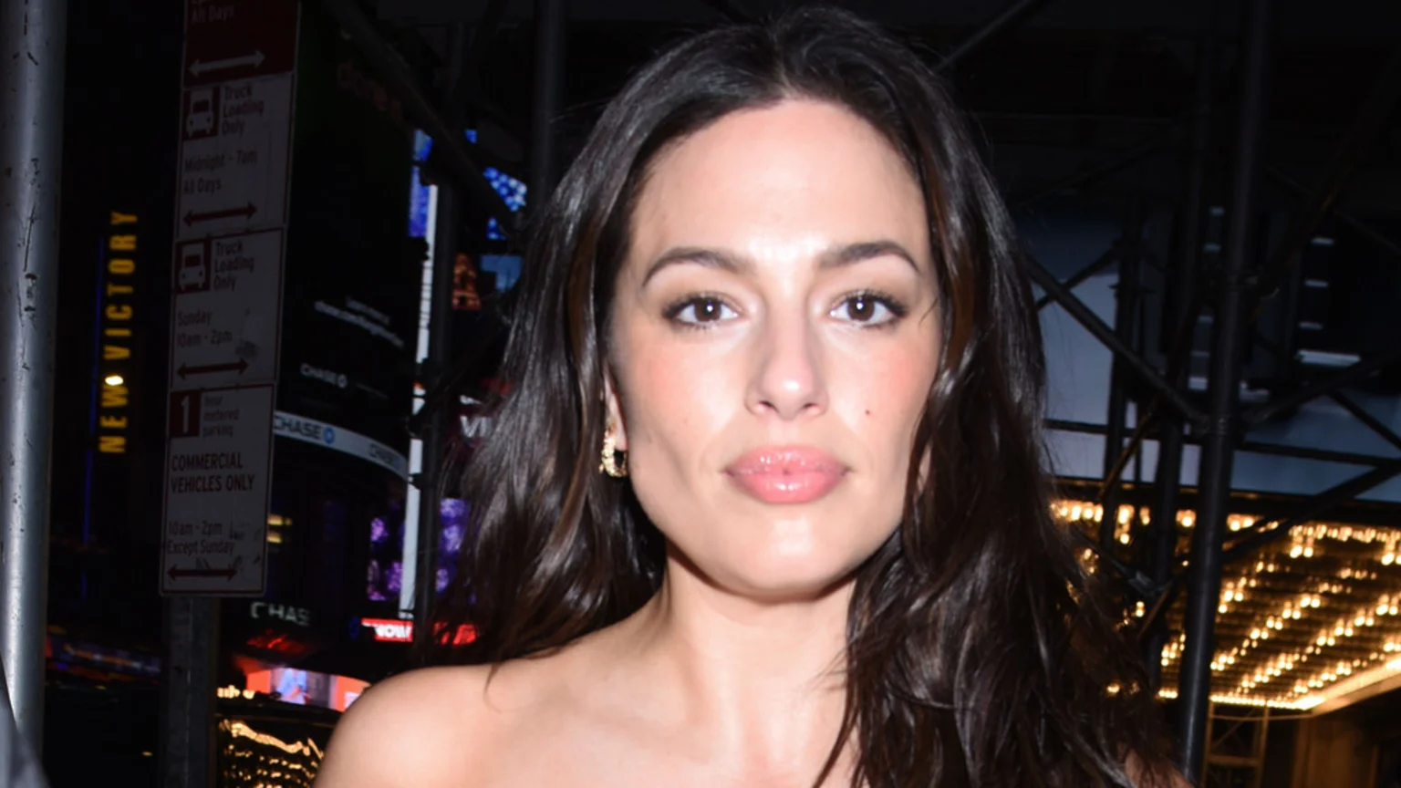 A close-up shot of model Ashley Graham with long, dark, wavy hair, light makeup, and glossy lips, wearing gold hoop earrings and a strapless top against a blurry nighttime city background.