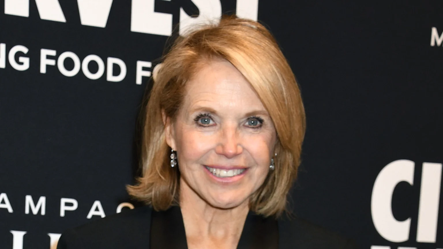 Katie Couric smiles brightly, featuring blonde hair, blue eyes, a black top, and dangling earrings, against a dark background with partially visible white text.