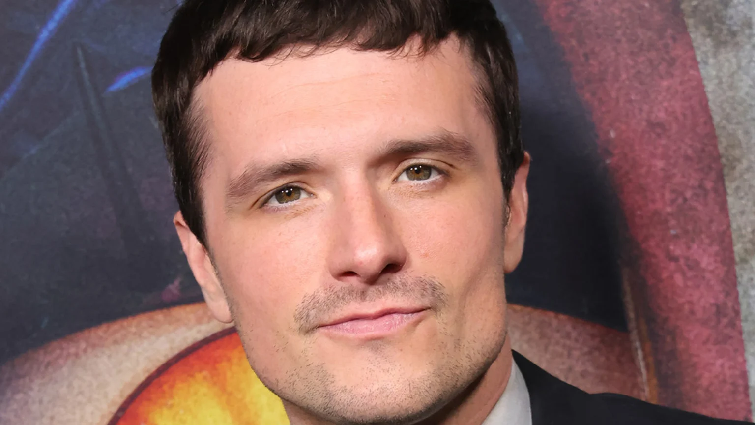 Close-up of actor Josh Hutcherson with short dark hair, light brown eyes, and light stubble, looking directly at the camera with a gentle smile while wearing a dark suit.