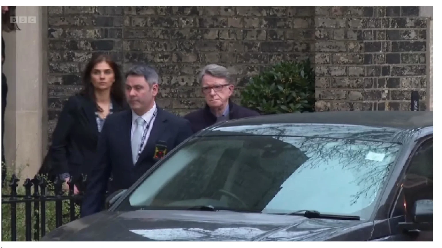 Three people, a man in a suit, a woman, and an older man in glasses, walk past a dark car and a black fence, with a brick building in the background.