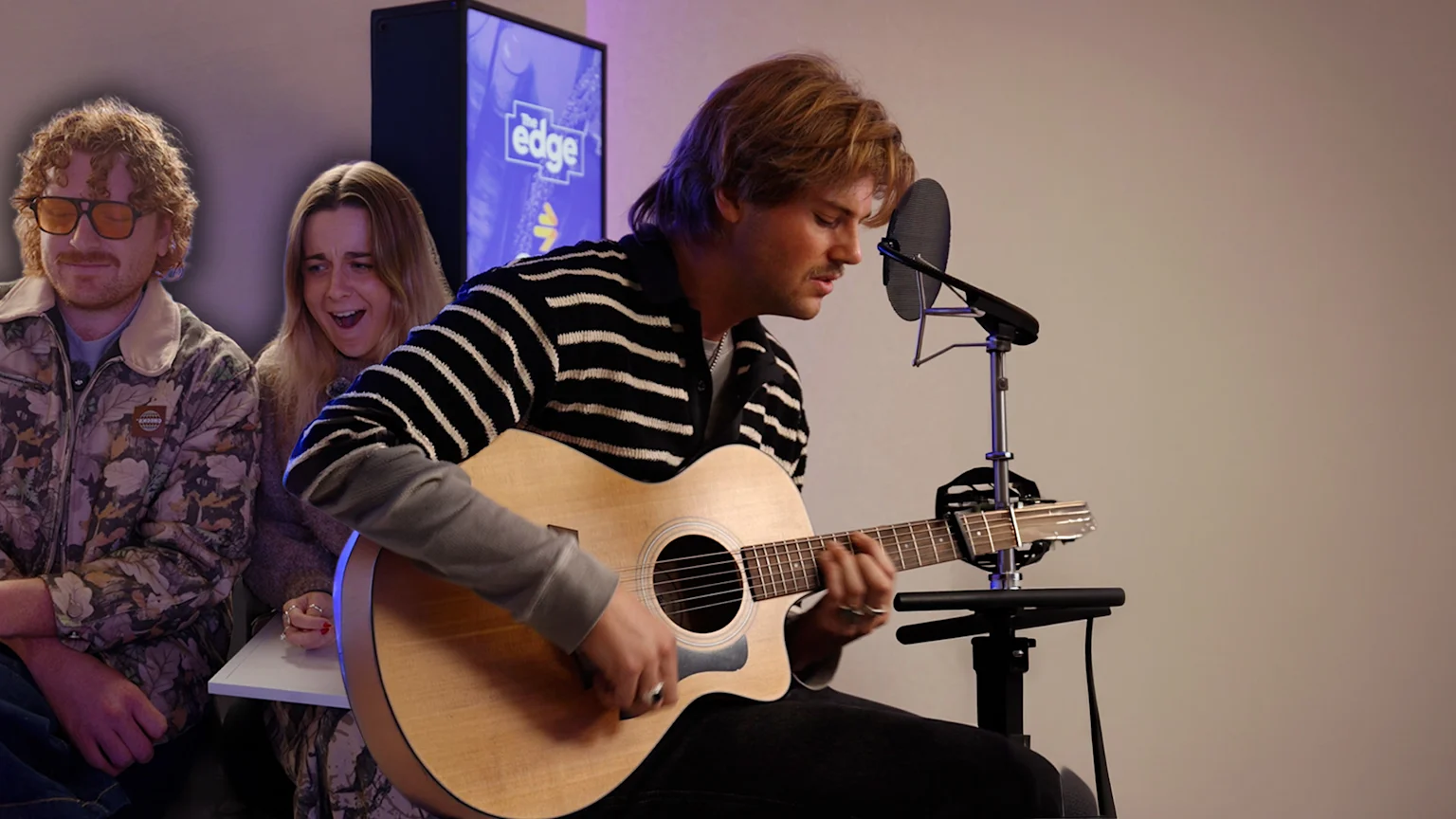 A musician in a striped sweater plays an acoustic guitar and sings into a microphone, while two people, a man in sunglasses and an excited woman, watch in the background near "The Edge" logo.