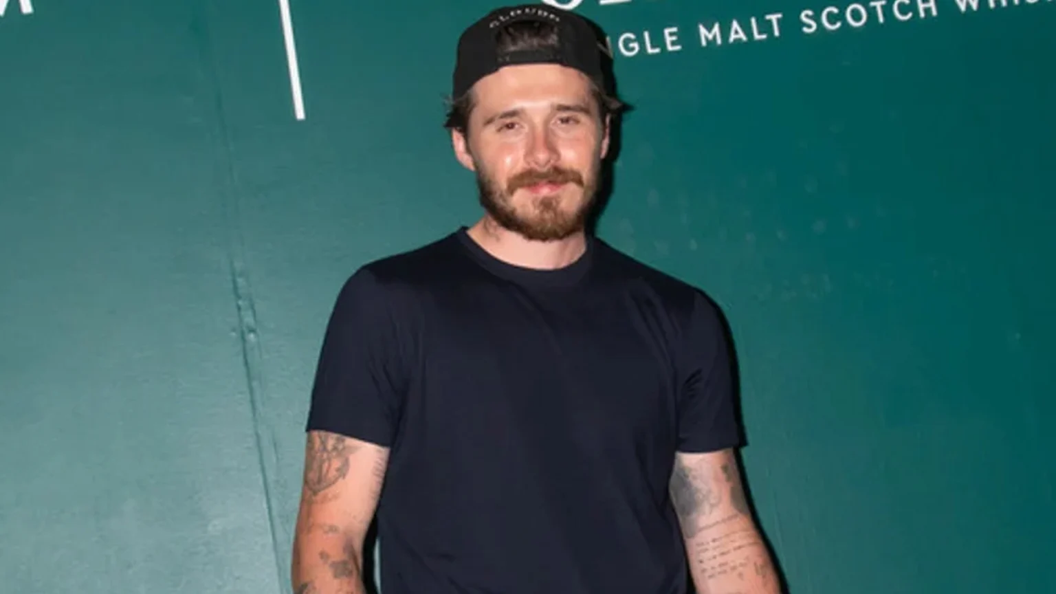 A man with a beard, tattoos, a black t-shirt, and a backward cap stands against a green wall featuring Aston Martin Formula One Team and Glenfiddich logos.