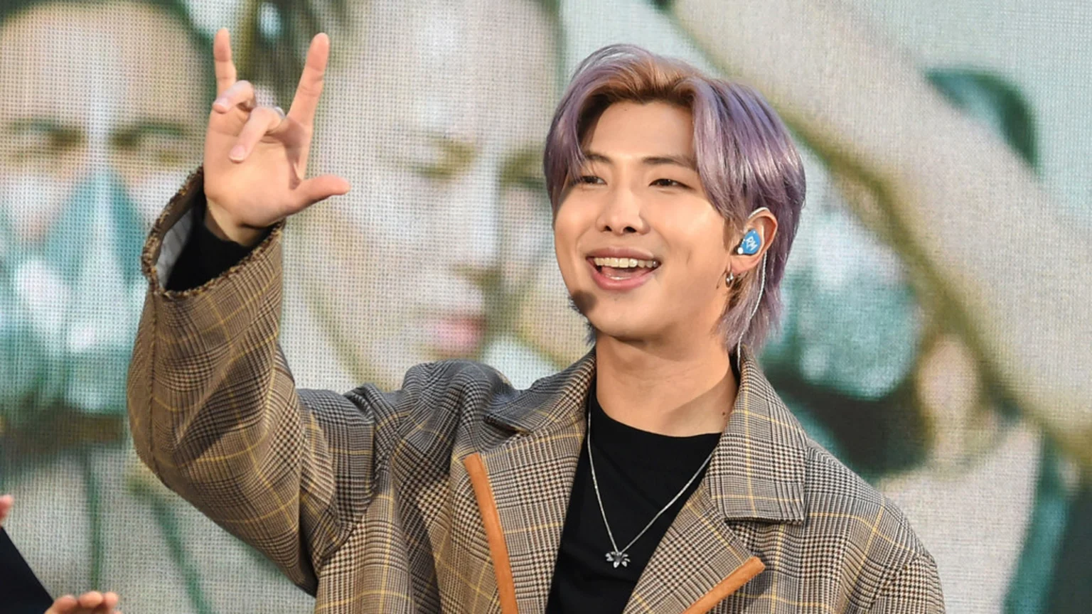 A man with purple hair smiles broadly, making a rock-and-roll hand gesture with his right hand, wearing a plaid jacket and a blue in-ear monitor with "RM" on it.