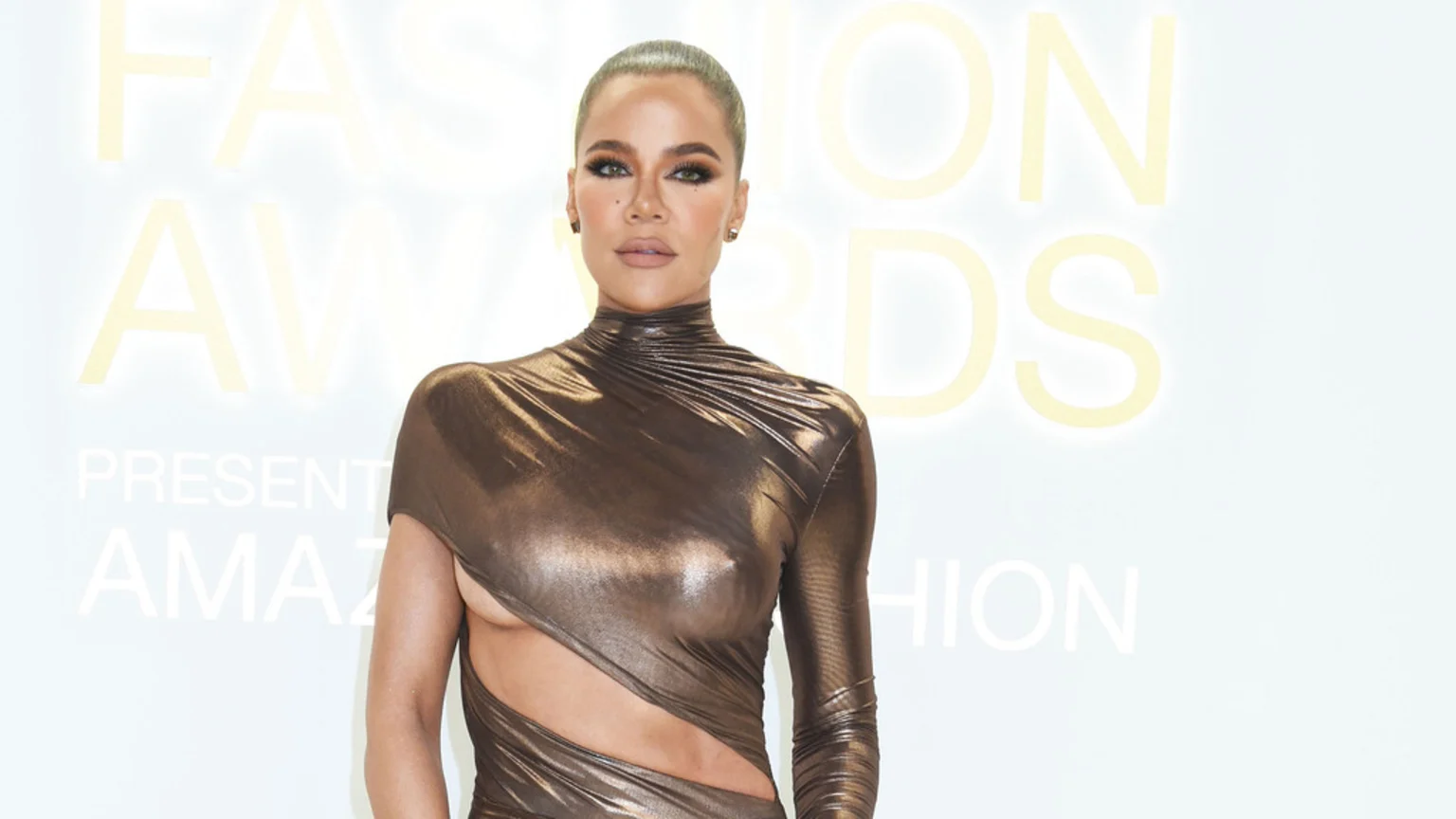 Khloé Kardashian poses in a shimmering bronze, high-neck dress with cut-outs, her hair pulled back into a bun, against a light background with faint text.