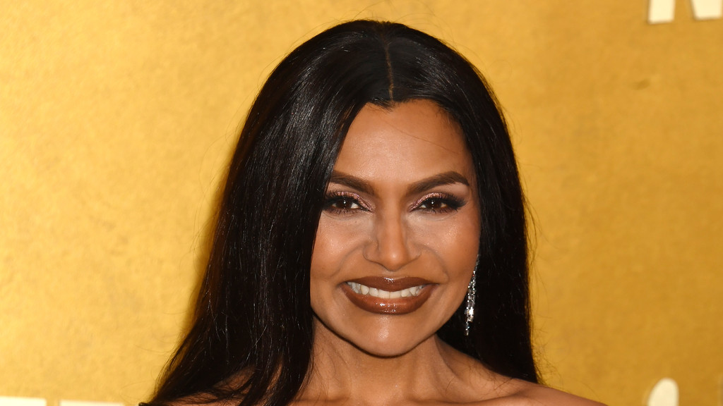 Close-up portrait of a smiling woman with long dark hair parted in the middle, wearing dark glossy lipstick, shimmery eyeshadow, and a dangle earring, against a yellow background.