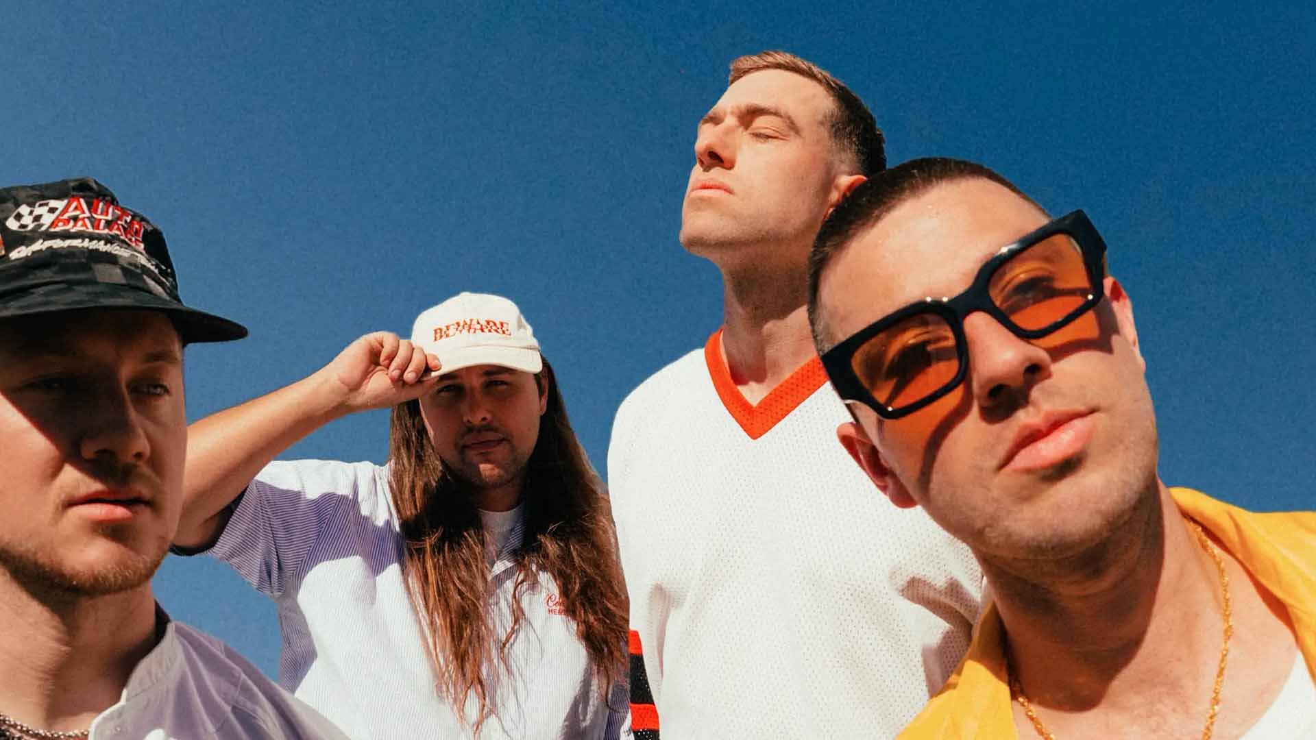 Four men pose under a clear blue sky; one wears a black bucket hat, another a "BEWARE" cap, a third a white V-neck shirt, and the fourth orange sunglasses.