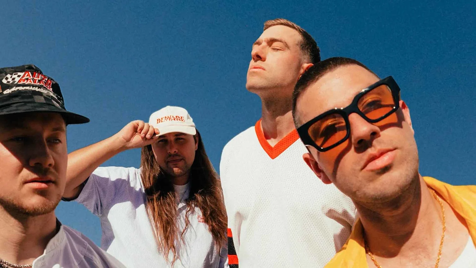 Four men pose under a clear blue sky; one wears a black bucket hat, another a "BEWARE" cap, a third a white V-neck shirt, and the fourth orange sunglasses.
