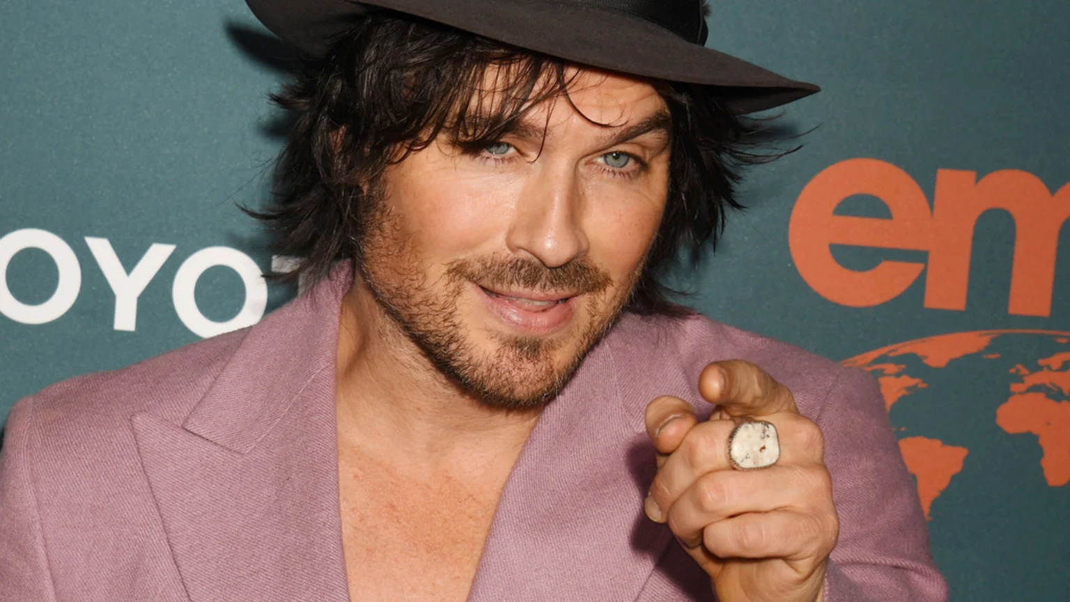 Ian Somerhalder, wearing a dark hat and pink jacket, with a beard and blue eyes, smiles and points directly forward at a branded event.