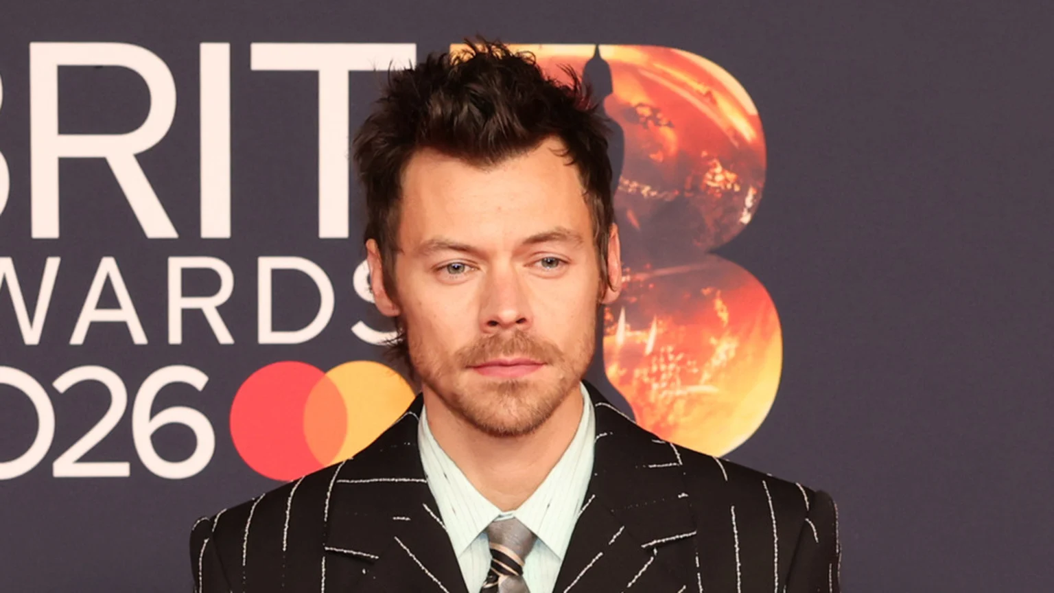 Harry Styles, with styled dark hair and a beard, looks directly at the camera wearing a pinstripe suit, light shirt, and striped tie, with "BRIT AWARDS 2026" and circular logos in the background.