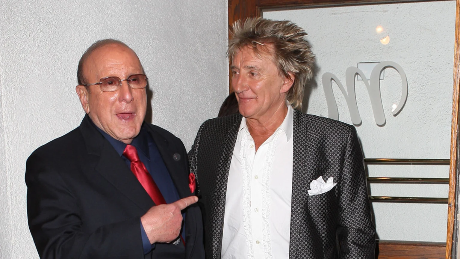 Clive Davis in a dark suit and red tie points at a smiling Rod Stewart in a patterned jacket and white ruffled shirt, standing in front of a white wall and glass door.