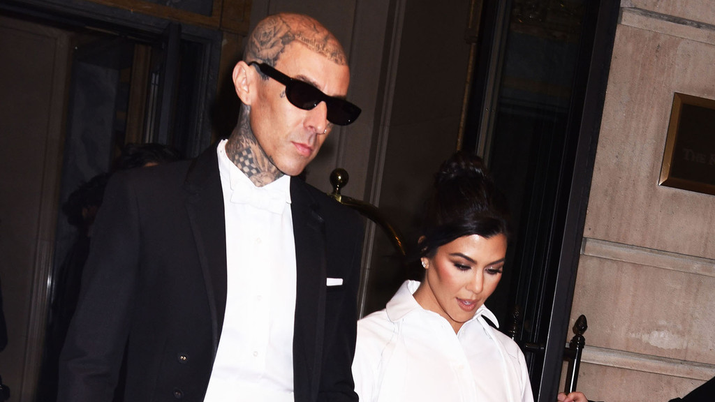 Travis Barker, bald with head tattoos and dark sunglasses, walks alongside Kourtney Kardashian, who has dark hair pulled into a bun and wears a white shirt.