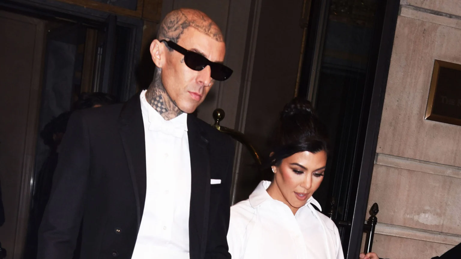 Travis Barker, bald with head tattoos and dark sunglasses, walks alongside Kourtney Kardashian, who has dark hair pulled into a bun and wears a white shirt.