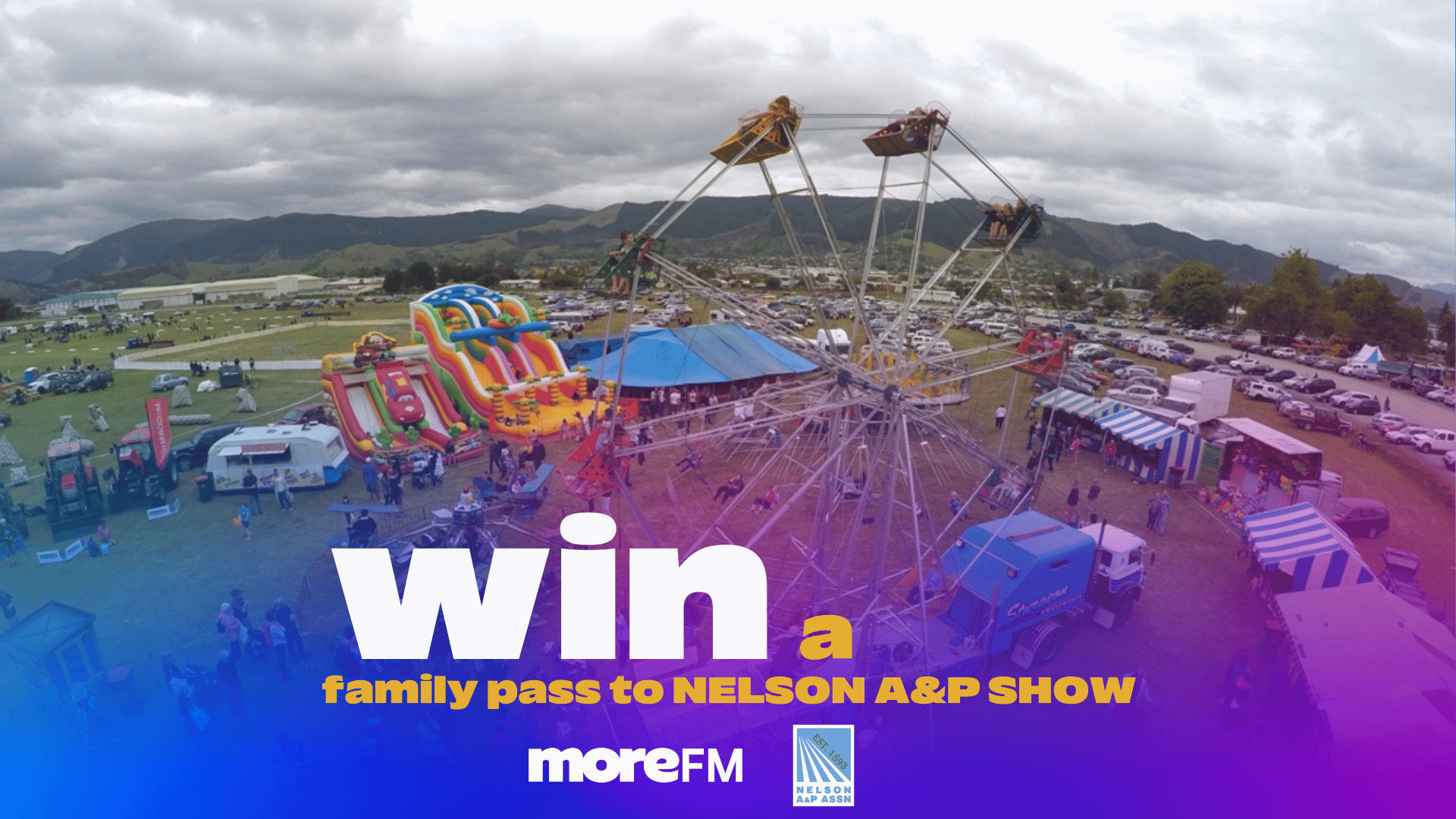 An aerial view of the Nelson A&P Show features a Ferris wheel, colorful inflatable slides, various tents, and crowds of people enjoying the outdoor event on a cloudy day, with mountains in the background.