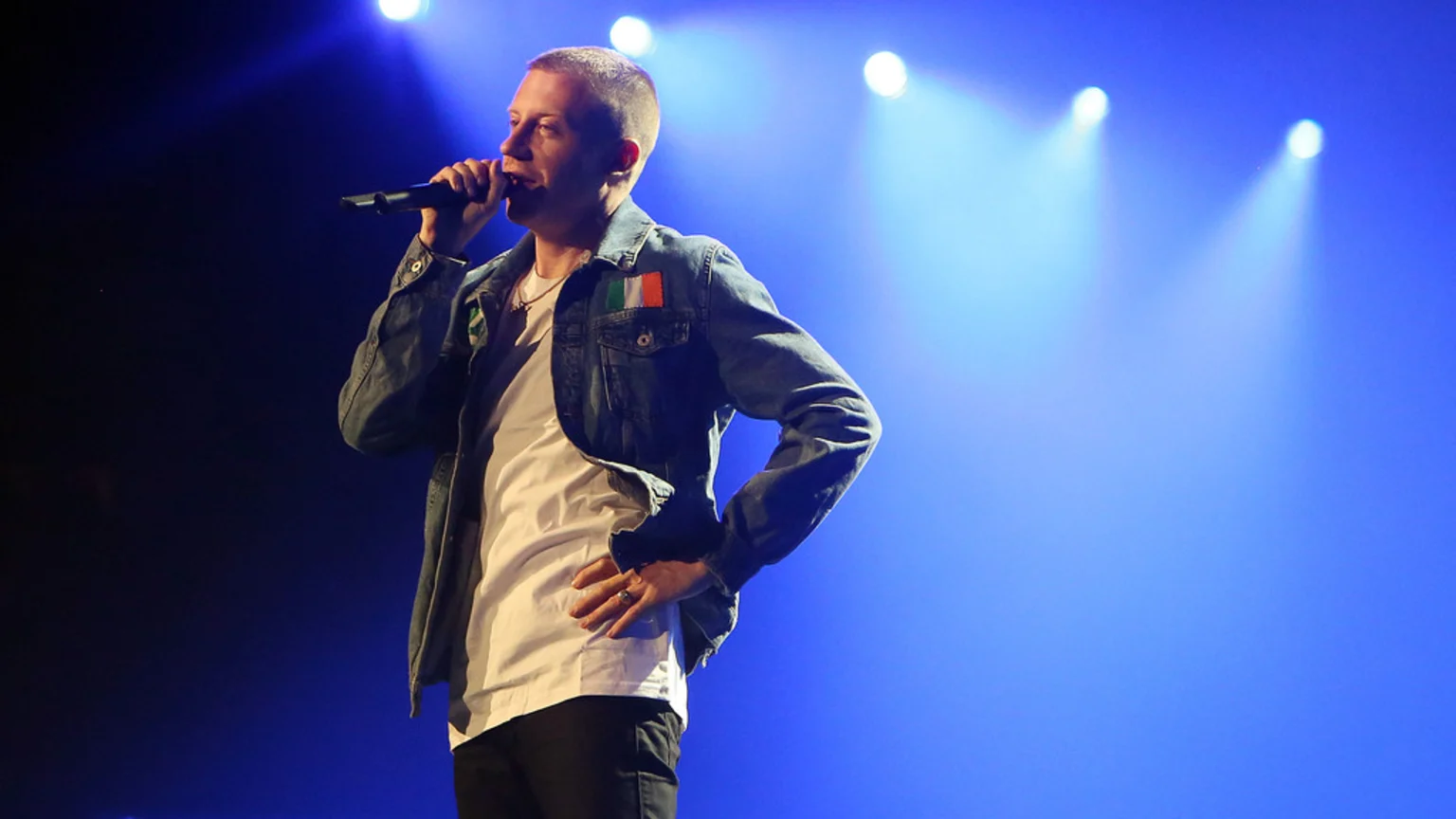 Macklemore performs on stage, holding a microphone, wearing a denim jacket with an Irish flag patch over a white t-shirt, with blue stage lights in the background.