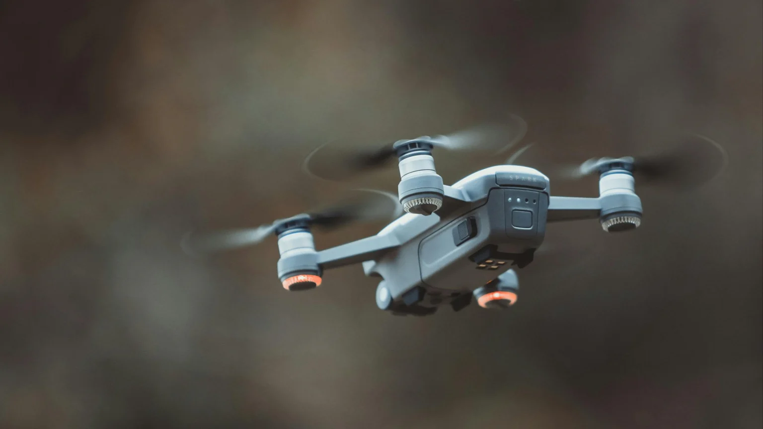 A grey DJI Spark drone with spinning propellers and glowing orange lights hovers in mid-air against a blurred, dark background.