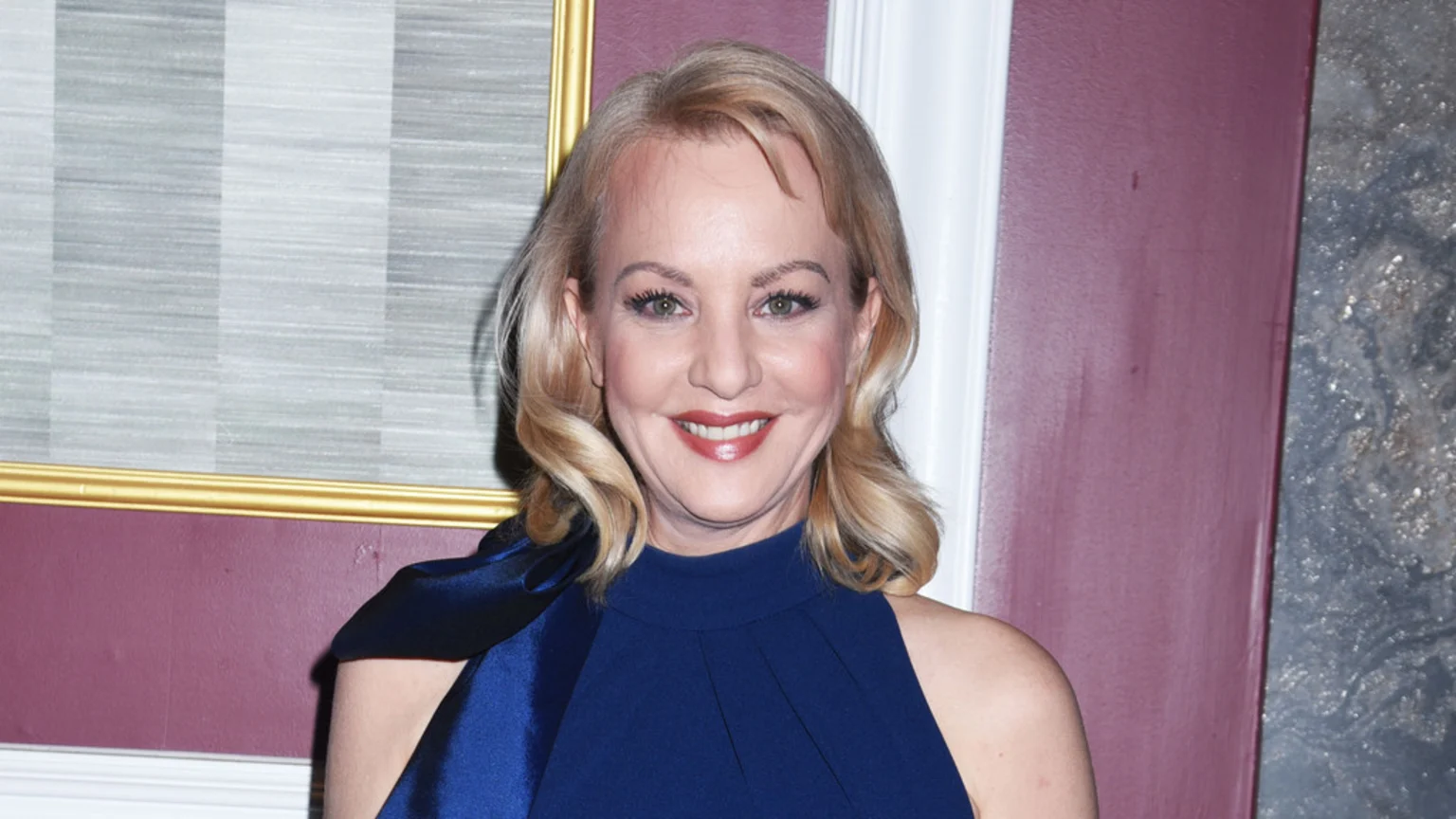 Actress Wendi McLendon-Covey, a blonde woman in a dark blue dress, smiles broadly at the camera in front of a patterned wall with a gold frame.