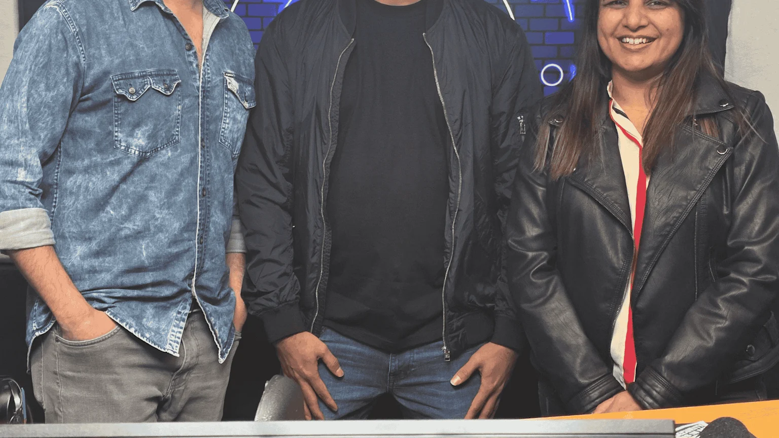 Three people are shown: a man in a denim shirt, a man in a black bomber jacket, and a smiling woman in a black leather jacket, against a dark background.