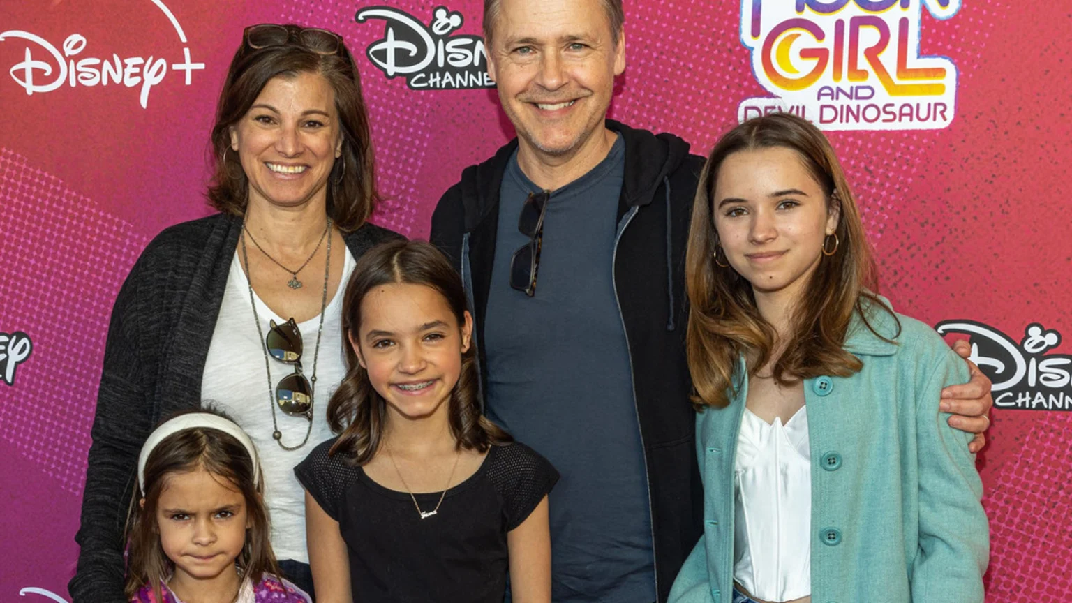 A family of five, including a man, a woman, and three girls, smile while posing in front of a colorful Disney event backdrop.