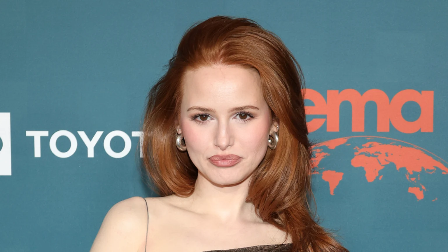 Close-up portrait of Madelaine Petsch with long red hair, wearing a dark thin-strap top and large metallic hoop earrings. She poses against a teal event backdrop featuring white Toyota and orange EMA logos with a world map graphic, under studio lighting.