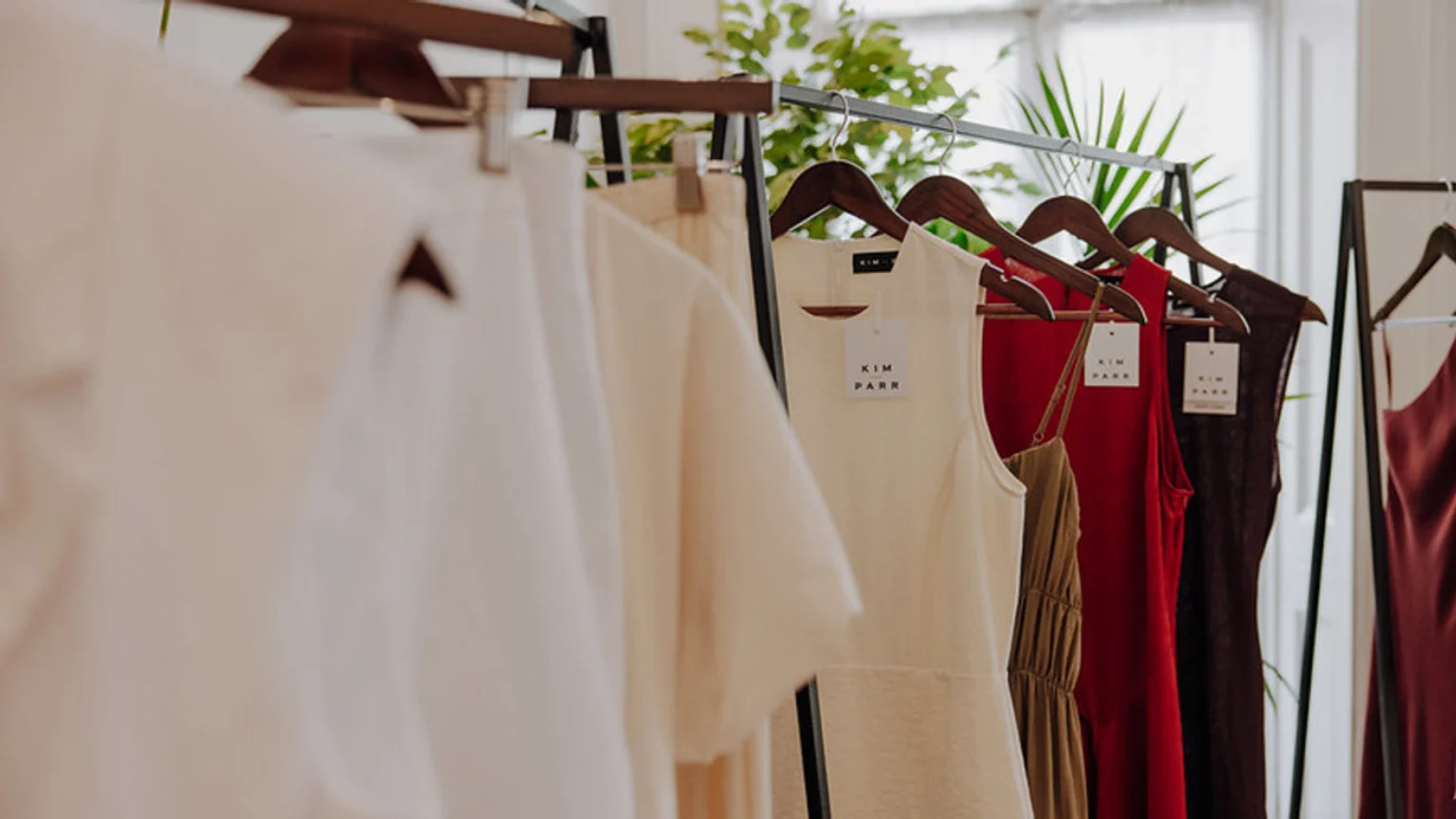 A clothing rack displays various garments, including cream, red, and brown tops and dresses, some with "KIM PARR" tags, in a well-lit setting.