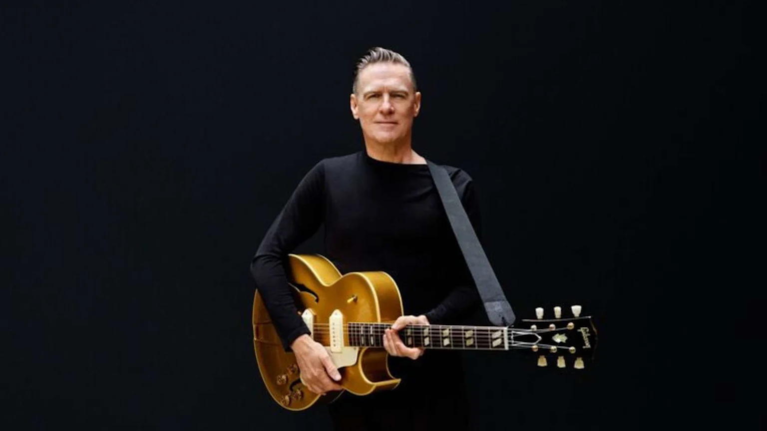 Musician Bryan Adams in a black shirt poses with a golden archtop guitar, looking forward against a dark background.